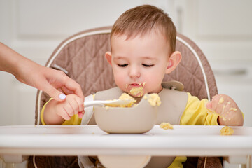 Mother teaches toddler baby boy to eat porridge with a spoon while sitting in a child chair. Mom...