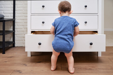 Toddler baby opens a chest of drawers. Child boy reaches into an open drawer of a white cabinet. Kid age one year - obrazy, fototapety, plakaty