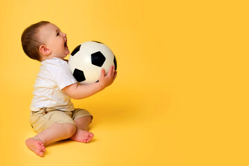 Happy baby boy plays with a soccer ball on a yellow studio background, copy space. A smiling child...