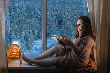 Young woman reading a book by the window in the light of the evening lamp. Woman sitting on the...