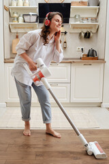 Smiling woman listens to music with headphones while dusting with a wireless portable vacuum...