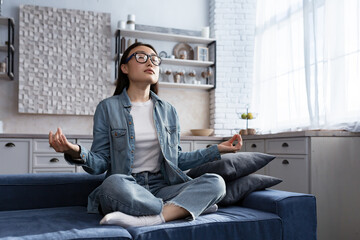 Fototapeta premium Beautiful Asian woman relaxing at home sitting on sofa and meditating in lotus pose, doing breathing exercises in living room