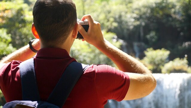 Wanderlust, tourism concept. Happy male tourist with backpack in travel. Young man traveler make photos of beautiful exotic waterfalls and tropical nature on retro style camera. Close-up, back view.