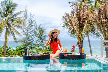 Happy woman in red swimsuit and straw hat with floating breakfast trays in luxury pool on villa...