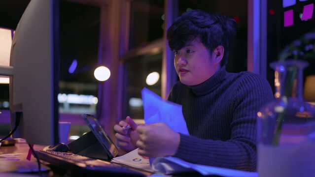 young Asia freelance creative designer programer focus working hand using pen with tablet thinking concentrate working late at home overtime at night low light ambient in living room with city bokeh