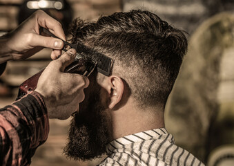 Hipster client getting haircut. Hands of barber with hair clipper, close up. Bearded man in barbershop. Haircut concept. Man visiting hairstylist in barbershop