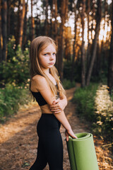 A beautiful preschool girl, a professional athlete in a black suit stands, turning around with a green rug in her hands for gymnastics and yoga in the forest at sunset. Photography, concept.