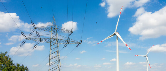 High voltage power lines and electricity transmission tower against blue sky with wind turbine on...