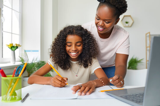 Help with homework. Joyful african american teenage girl doing school homework at home with mom's help. Smiling young dark-skinned woman watches her daughter write in notebook. Education and family.