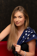 a beautiful blonde 20-25 years old with long hair holds an American flag in her hand on a black background, sitting on a red sofa.