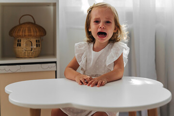 A little girl cries and is naughty sitting at the table in the nursery. Children's tantrums, tears...