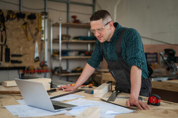 Portrait of a carpenter in protective glasses and work overalls uses a laptop in a workshop.