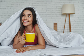Dreamful woman looking out of a blanket and holding coffee cup while lying in bed at home - Powered by Adobe