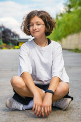 Portrait of caucasian teenage girl with curly hair in cool trendy glasses sitting on asphalt in city park on sunny summer day