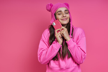 Obraz na płótnie Canvas Joyful young woman in funky hat holding smart phone near face against pink background