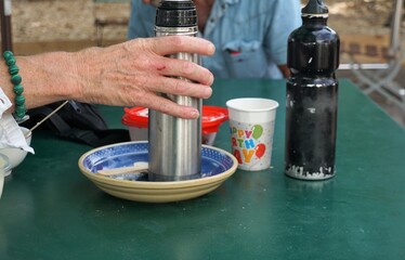 Frauenhand umfasst silberne Kaffeekanne aus Metall über Tisch mit Teller, buntem Pappbecher und schwarzer Flasche auf grüner Metalltischplatte vor Mann mit blauem Jeanshemd am Nachmittag im Sommer