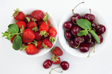 Two white plates with cherries and strawberries with mint petals stand on a white background. High quality photo
