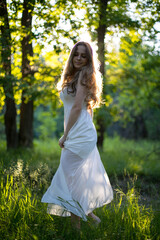 Beautiful young Russian girl dressed in a white dress, walks through the forest among the oaks in the rays of the sunset