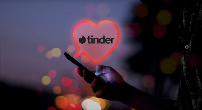 Tinder logo in an illustrated heart shaped speech bubble above a hand holding a smartphone with screen glowing at night, red, orange and yellow dots of light, romantic, dating, flirt, love