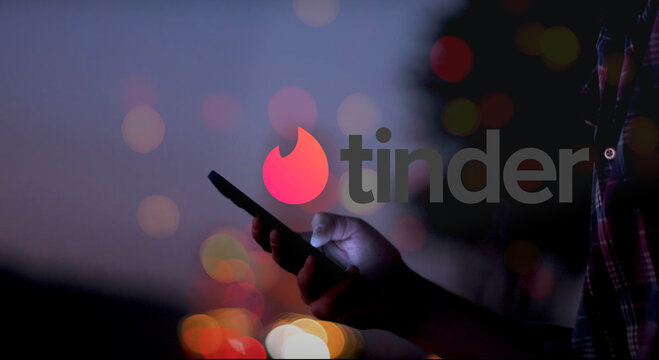 Tinder Logo over a hand holding a smartphone with glowing screen at night, red, orange, and yellow light dots in the background, romantic, dating, flirt, love