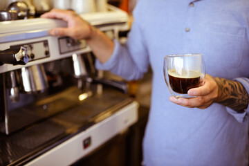 Faceless shot of tattooed barista in shirt holding glass of coffee near blurred machine in cafe 