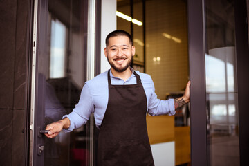Positive asian owner in apron and shirt looking at camera near entrance of cafe 