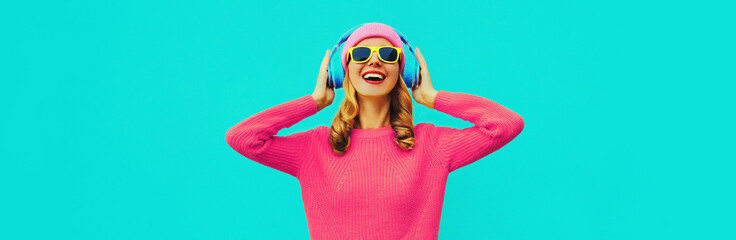 Portrait of happy smiling young woman with headphones listening to music wearing colorful pink sweater on blue background