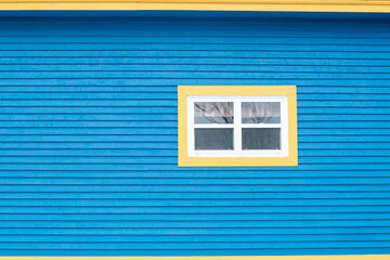 The exterior wall of a wooden v groove clapboard siding house with narrow vertical boards. There's a small closed glass window with four panes and white and yellow trim. A white lace curtain hanging 