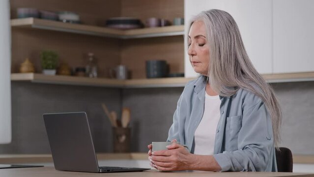 Caucasian businesswoman housewife senior freelancer mature lady sitting at home kitchen holds cup of tea or hot coffee in hands keeps warm works on laptop chatting online turns head at camera smiling