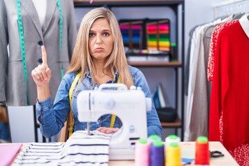 Blonde woman dressmaker designer using sew machine pointing up looking sad and upset, indicating direction with fingers, unhappy and depressed.
