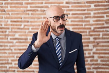 Bald man with beard wearing business clothes and glasses smiling with hand over ear listening an hearing to rumor or gossip. deafness concept.