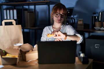 Young beautiful woman working using computer laptop and eating delivery food checking the time on wrist watch, relaxed and confident