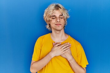 Young modern man standing over blue background smiling with hands on chest with closed eyes and grateful gesture on face. health concept.