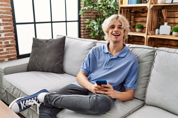 Young blond man using smartphone sitting on sofa at home