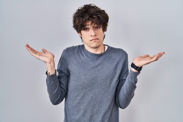 Young man standing over isolated background clueless and confused expression with arms and hands...