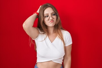 Young caucasian woman standing over red background confuse and wondering about question. uncertain with doubt, thinking with hand on head. pensive concept.