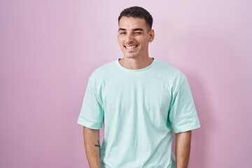 Handsome hispanic man standing over pink background winking looking at the camera with sexy expression, cheerful and happy face.