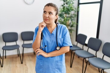 Young brunette doctor woman at waiting room serious face thinking about question with hand on chin,...