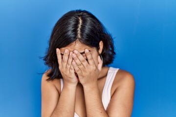 Young hispanic woman standing over blue background with sad expression covering face with hands while crying. depression concept.