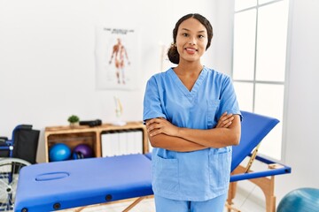 Young latin woman wearing physiotherapist uniform standing with arms crossed gesture at clinic