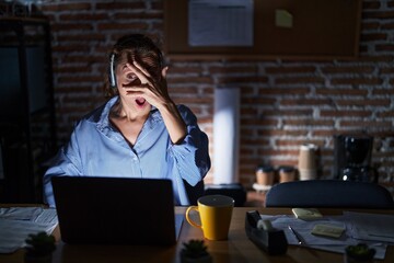 Beautiful brunette woman working at the office at night peeking in shock covering face and eyes with hand, looking through fingers with embarrassed expression.