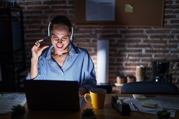 Beautiful brunette woman working at the office at night smiling and confident gesturing with hand doing small size sign with fingers looking and the camera. measure concept.