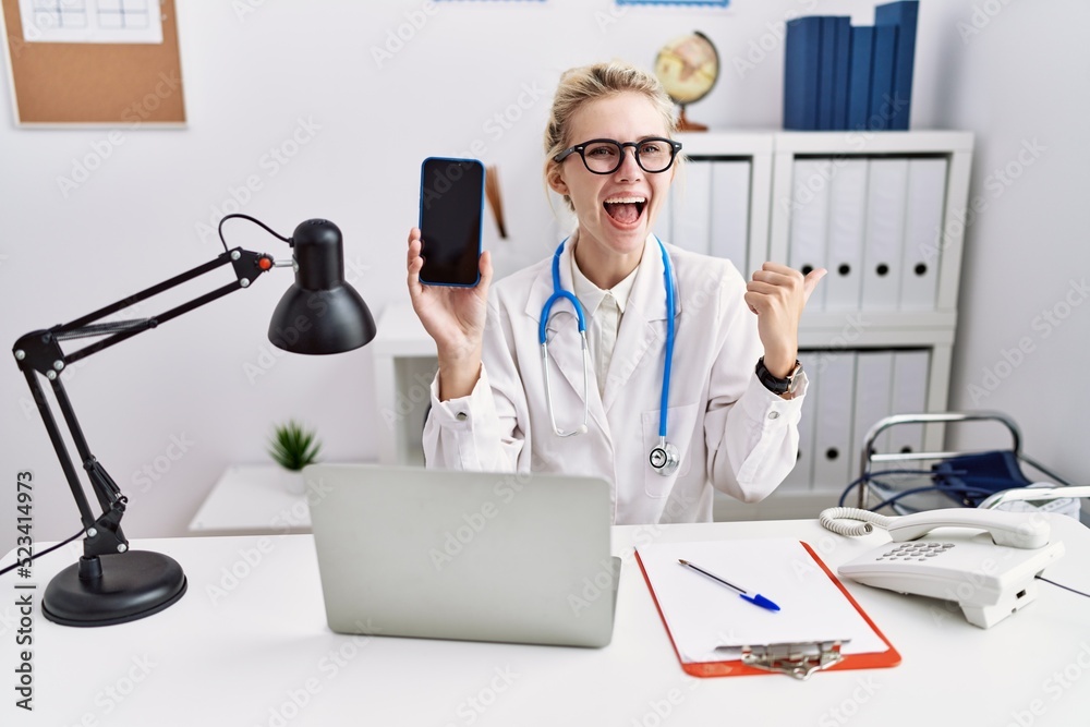 Sticker young doctor woman holding smartphone showing blank screen pointing thumb up to the side smiling hap