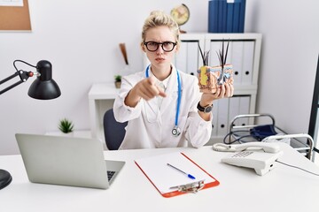 Young doctor woman holding model of human anatomical skin and hair at the clinic pointing with finger to the camera and to you, confident gesture looking serious
