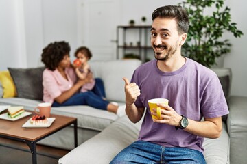 Hispanic father of interracial family drinking a cup coffee pointing to the back behind with hand and thumbs up, smiling confident