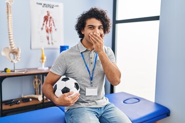 Hispanic man with curly hair working as football physiotherapist laughing and embarrassed giggle...
