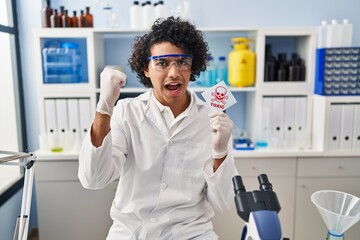 Hispanic man with curly hair working at scientist laboratory holding toxic banner annoyed and frustrated shouting with anger, yelling crazy with anger and hand raised