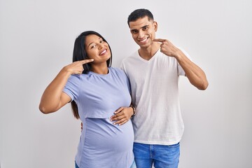 Young hispanic couple expecting a baby standing over background smiling cheerful showing and pointing with fingers teeth and mouth. dental health concept.