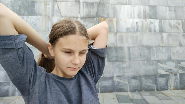 Cute teen girl gathers her blonde loose hair in ponytail. Brown long shaggy hair. Hair scrunchie. Young beautiful woman near stonewall. Casual hairstyle walk big city. Portrait girl without makeup