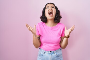 Young hispanic woman standing over pink background crazy and mad shouting and yelling with aggressive expression and arms raised. frustration concept.